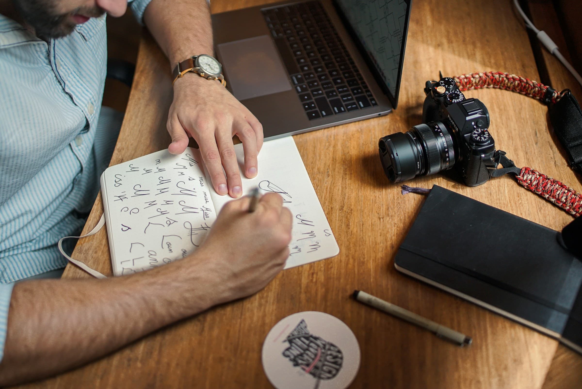 A designer's hand sketching lettering in a notebook on a warm wooden desk, wristwatch visible, DSLR and laptop in the frame.